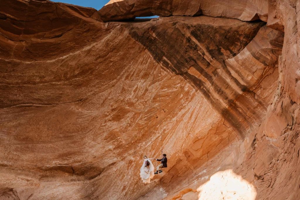 A couple repels down together from a hanging repel off of an arch in Moab in their wedding attire.