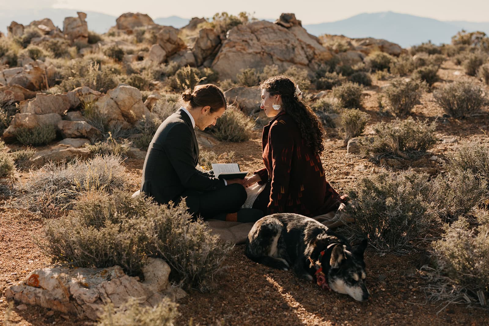 A couple sits together in the sun of the desert by their dog as the groom reads his vows.