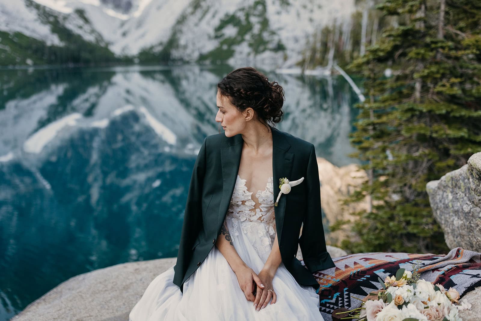 A photo of a bride sitting by a lake with her groom's jacket over her shoulders.