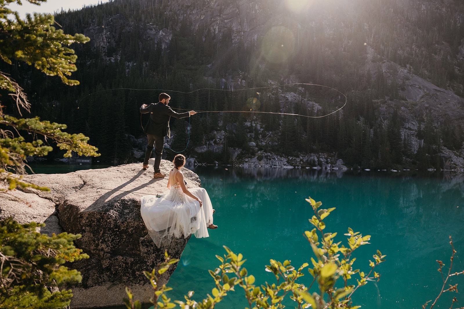 A bride and groom together on a rock atop a turquoise colored lake where the groom is fly fishing.
