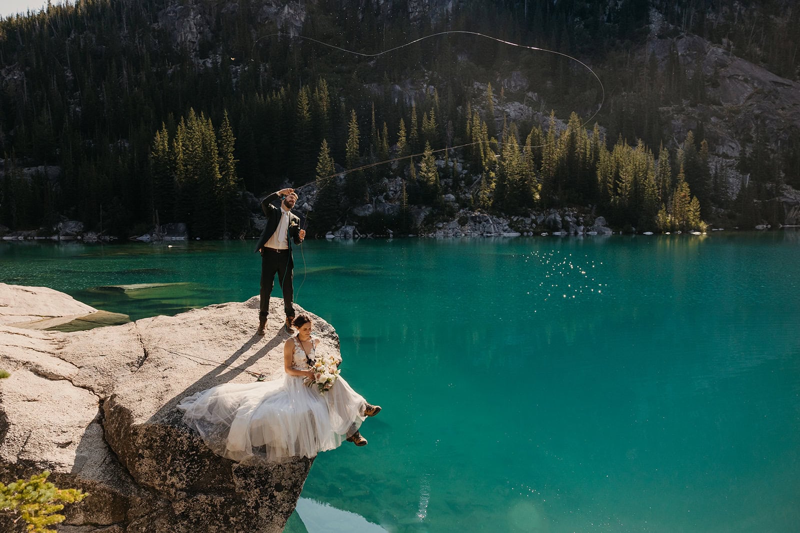 A bride and groom together on a rock atop a turquoise colored lake where the groom is fly fishing.