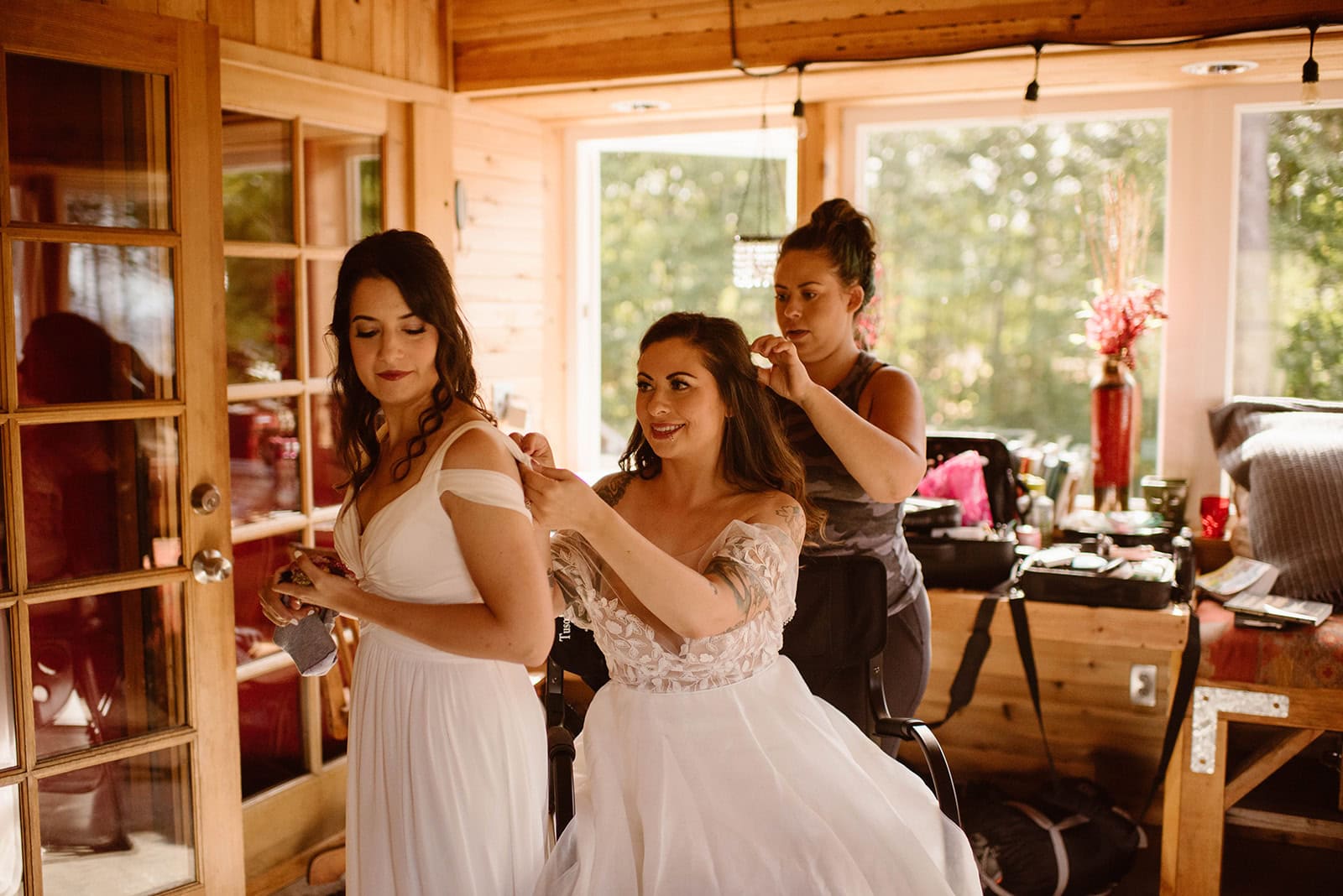 A make up artist does final touches while a bride helps her future wife with her dress.