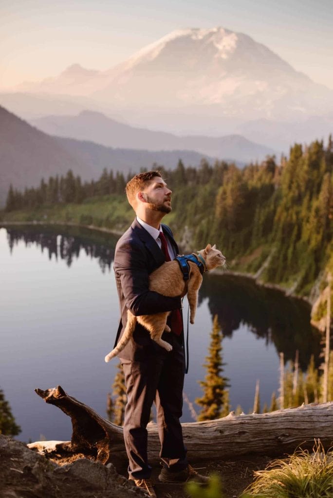 A groom holds his cat as they look at birds together with a mountain behind them.