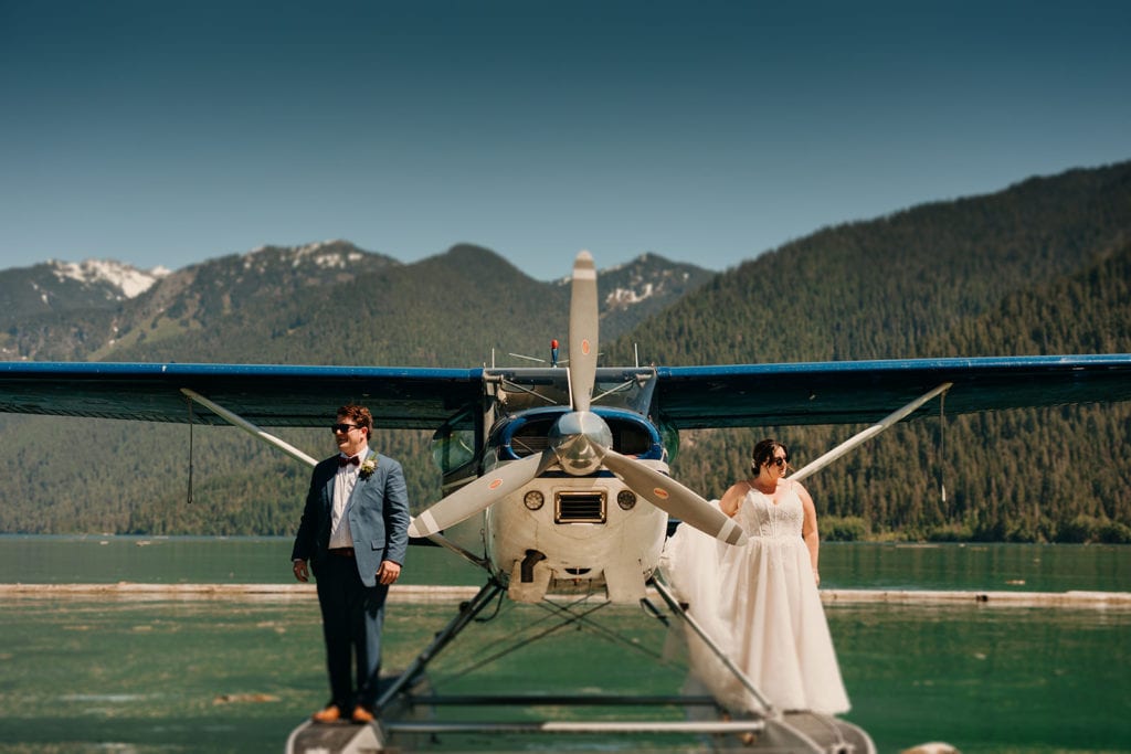 A couple stands on a seaplane on a lake in their wedding attire in the mountains.