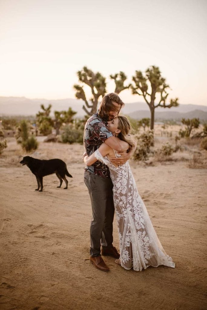 A bride and groom embrace while their dog stands nearby.