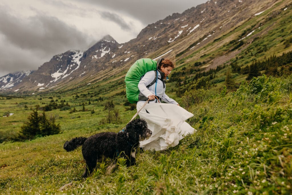 A bride hikes up a mountain on a cloudy alaska day with her dog following her.
