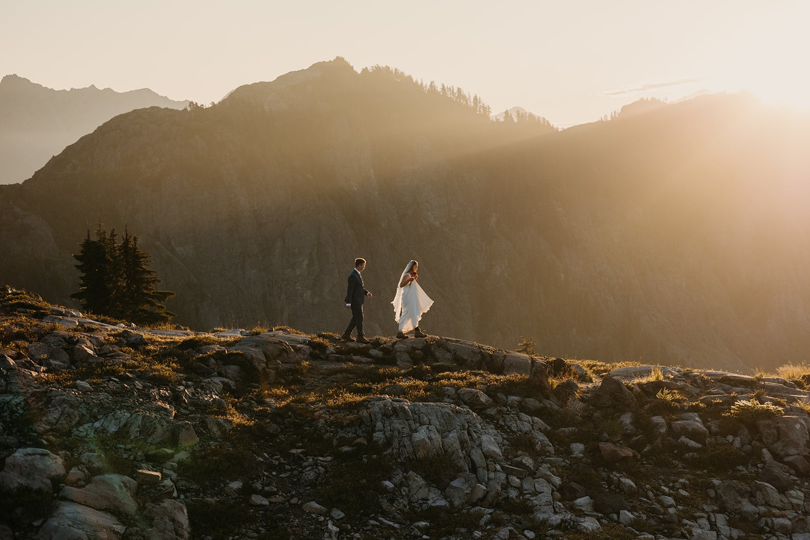 A bride and groom walk down the side of a mountain in the morning light.