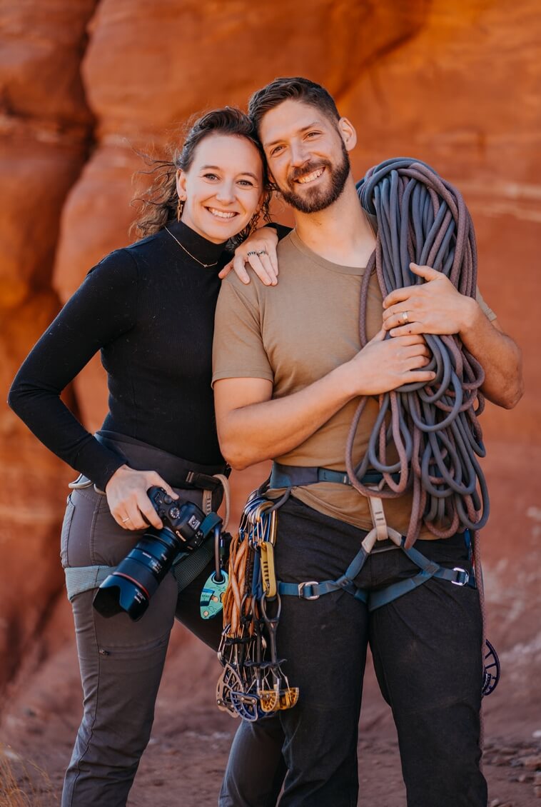A couple standing close together while one holds repelling gear and the other holds a camera