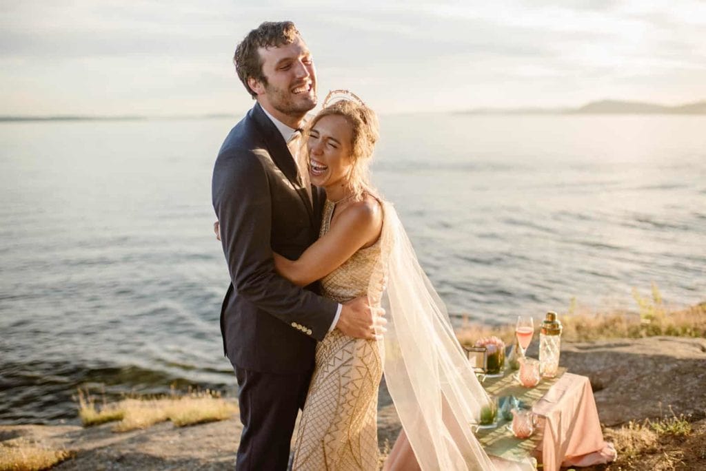 A bride and groom hugging each other in celebration at sunset right at the shore line.