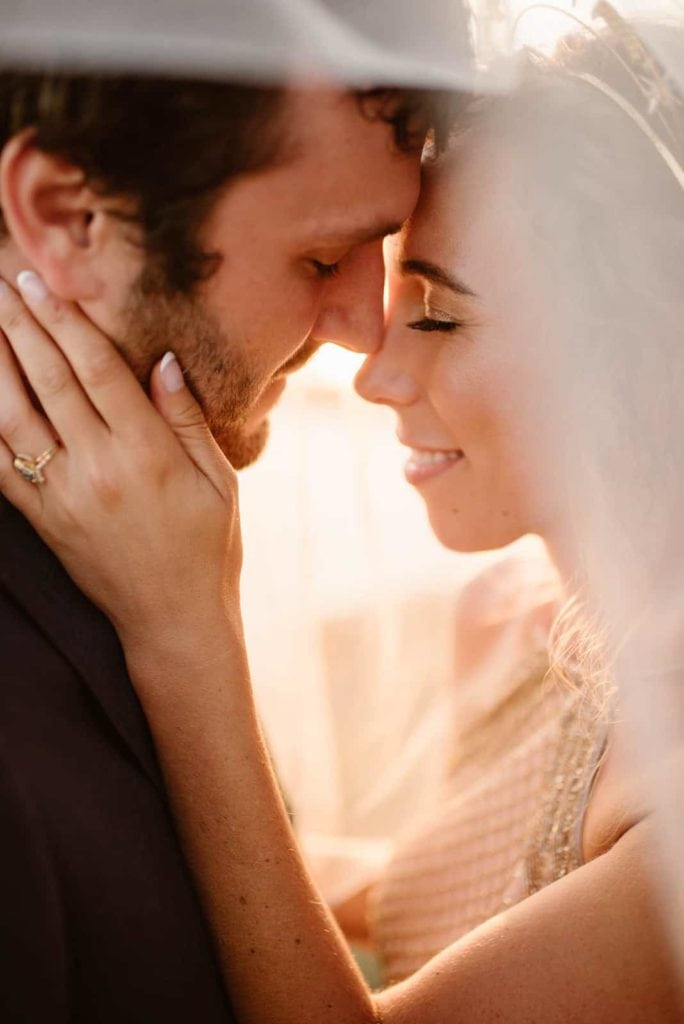 A close up photograph of a bride and groom standing with their foreheads touching and sharing a soft smile.