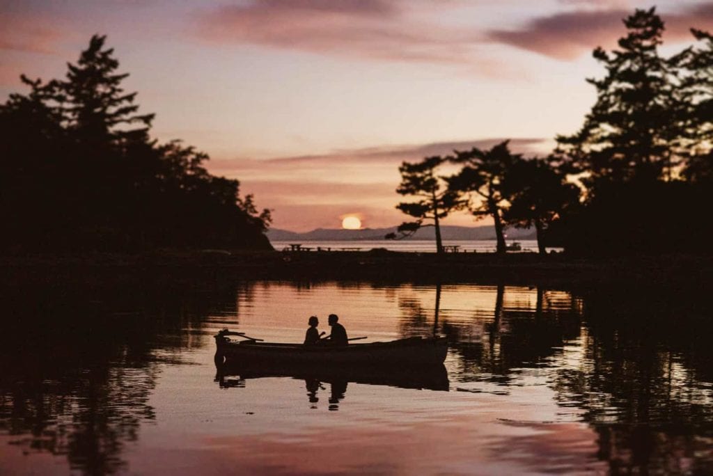A late sunset picture of a couple sitting together in a boat with pink, purple and blue colors in the sky.