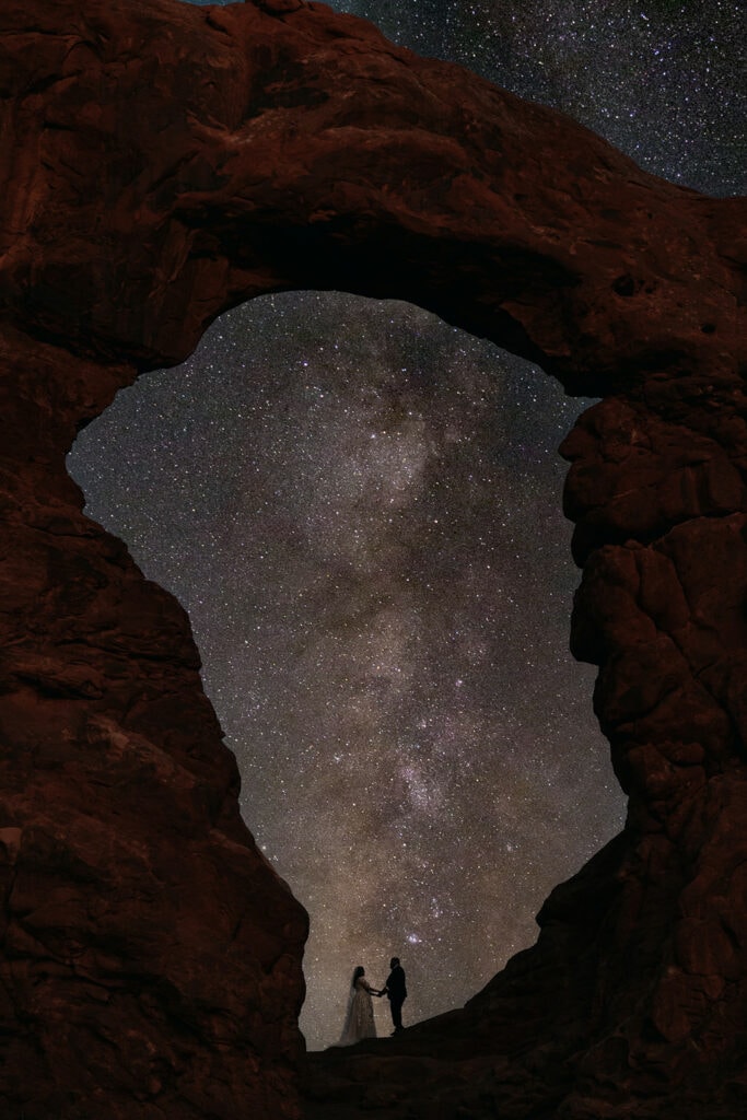 The couple stands under the arch under the milky way.