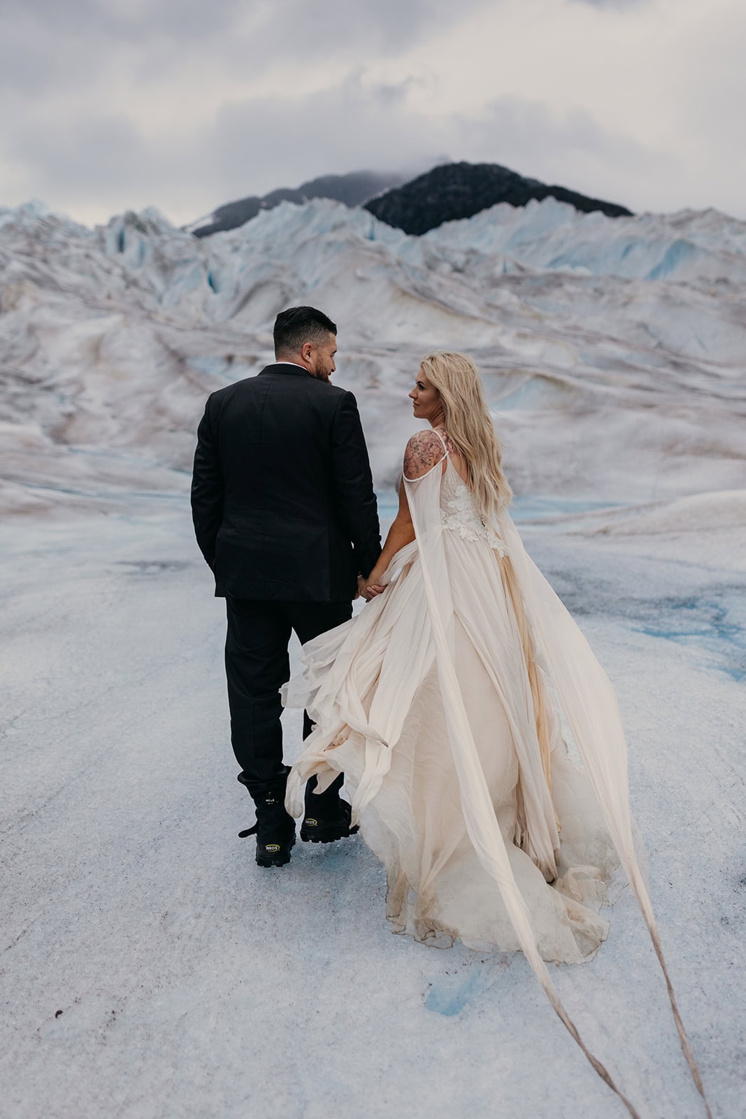 A bride and groom walk away from the camera on a glacier, the silk in her Daci gown blowing in the wind.