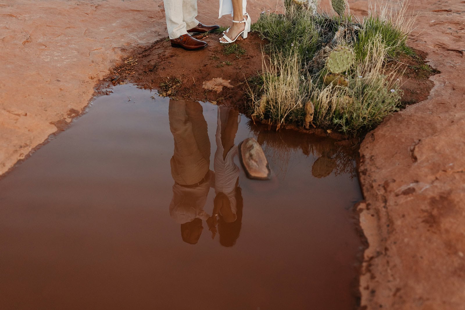 A detail photo of a couples shoes, bu t you can see the couple in the reflection of the still water. 