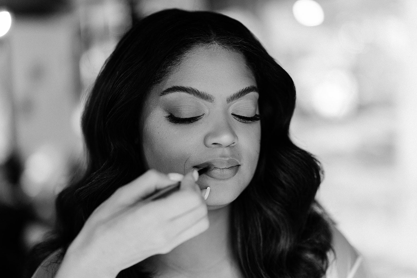 A bride sits by a window as the hair and makeup artist does a final touch up on her lips.
