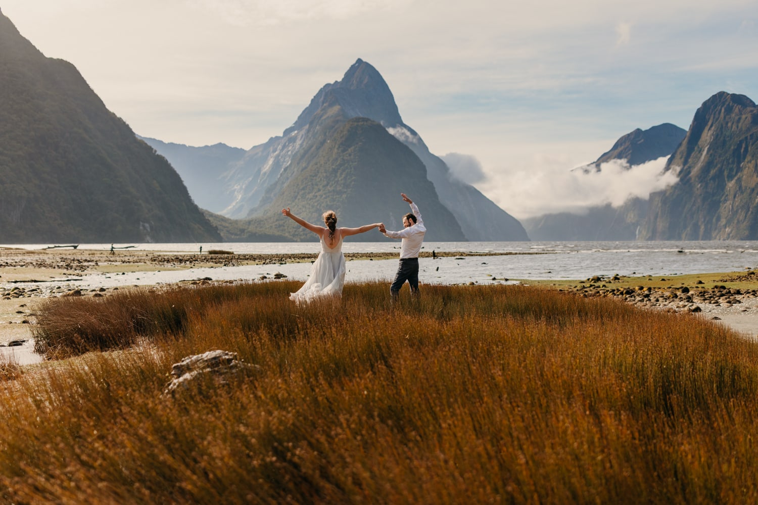 A bride and groom raise their hands in celebration at milford sound.
