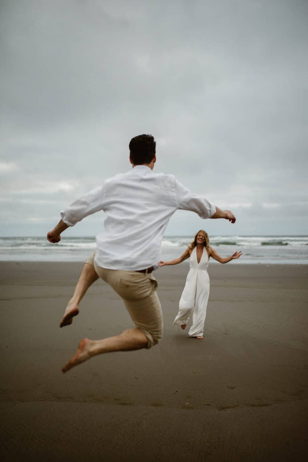 A groom jumps for joy on the beach running towards his bride.
