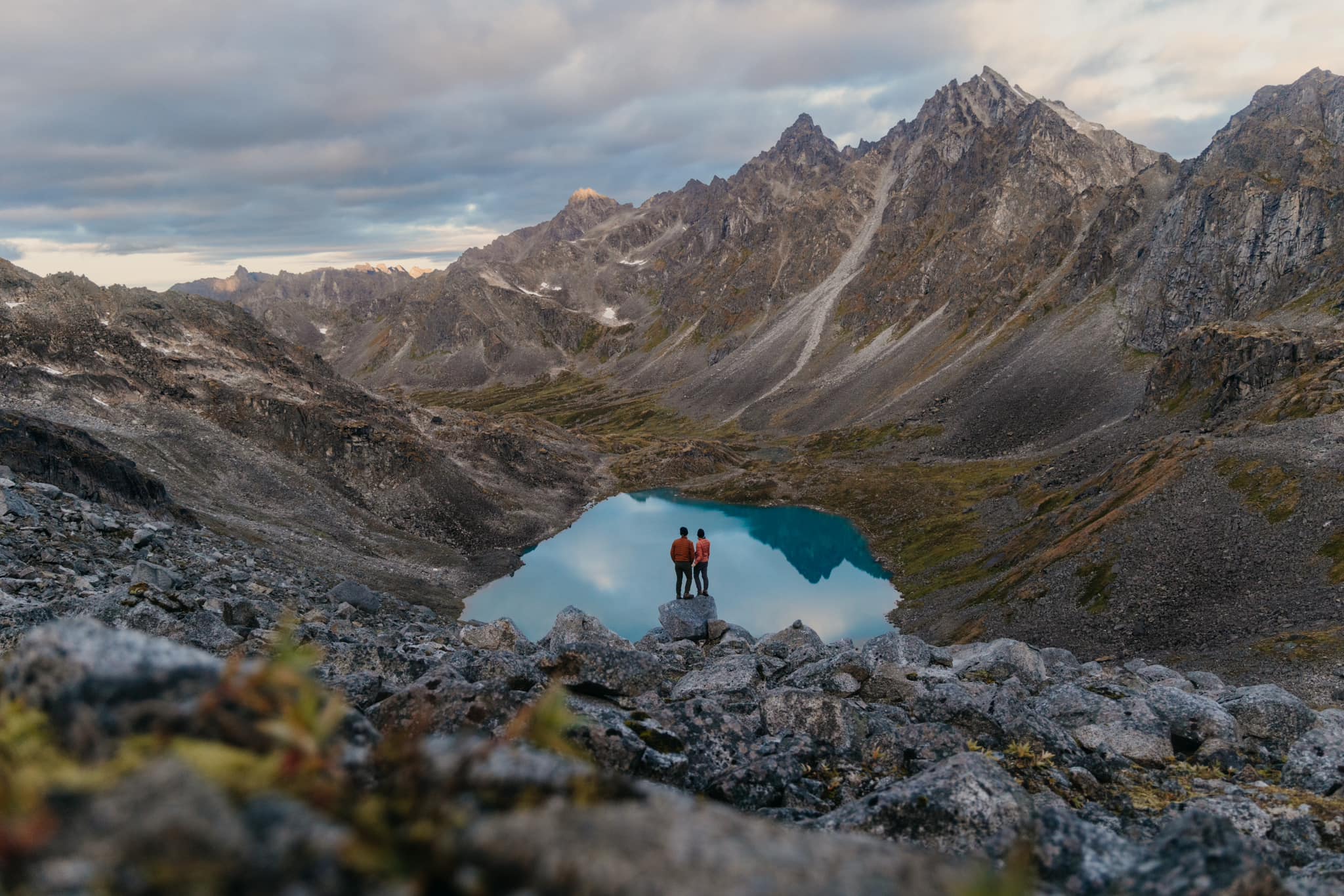 A couple of photographers stand on a rock overlooking big mountains and an alpine lake in Alaska.