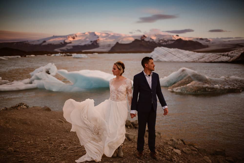 A bride and groom holds hands looking opposite directions standing at a lagoon in Iceland at midnight under the summer solistice.