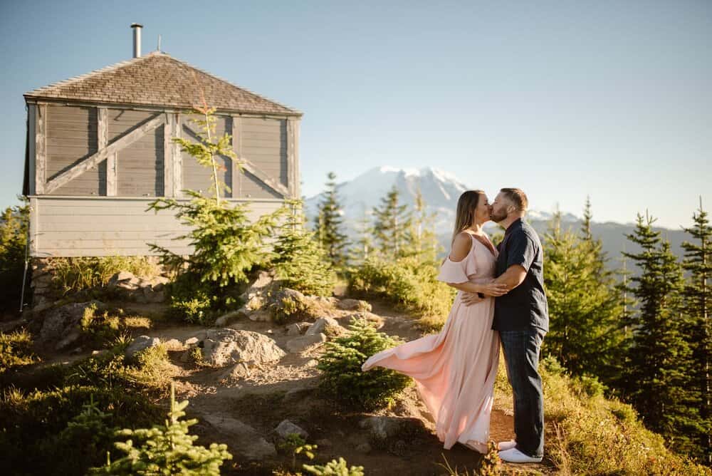 A couple shares a kiss with Mt Rainier and a fire tower in the background. 