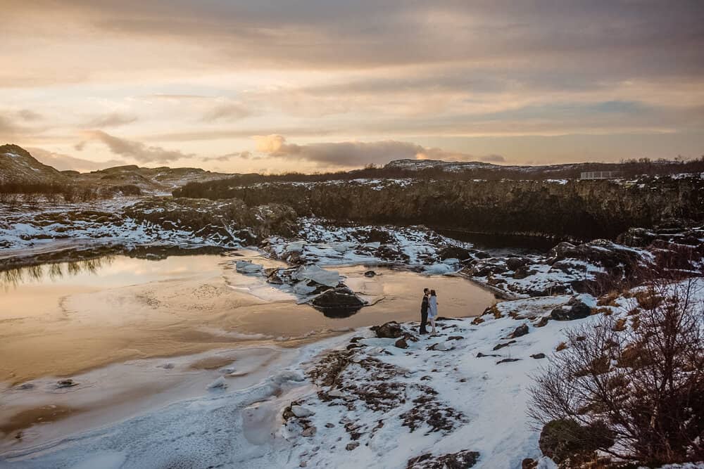 A couple stands by a body of water surrounded by snow on a Winter day in Iceland on the morning of their elopement.
