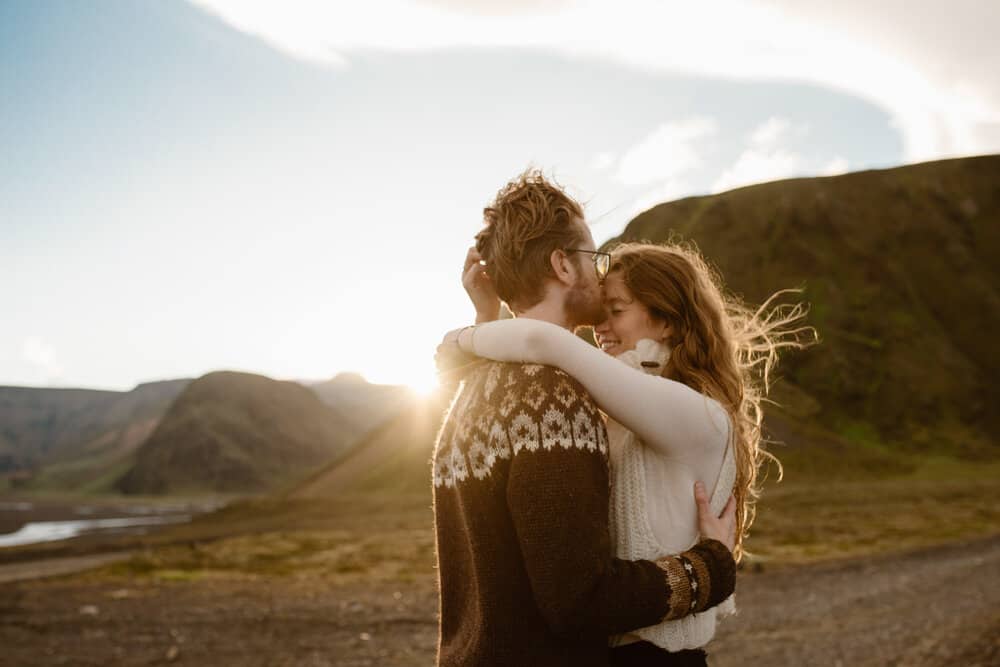 A man kisses a woman on the forehead while the sun sets below the horizon in the distant background.