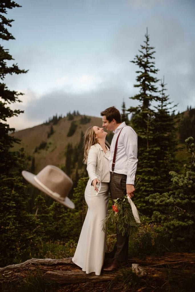 A bride throws her hat at the camera as she smiles at their husband while standing on a log in the forest.