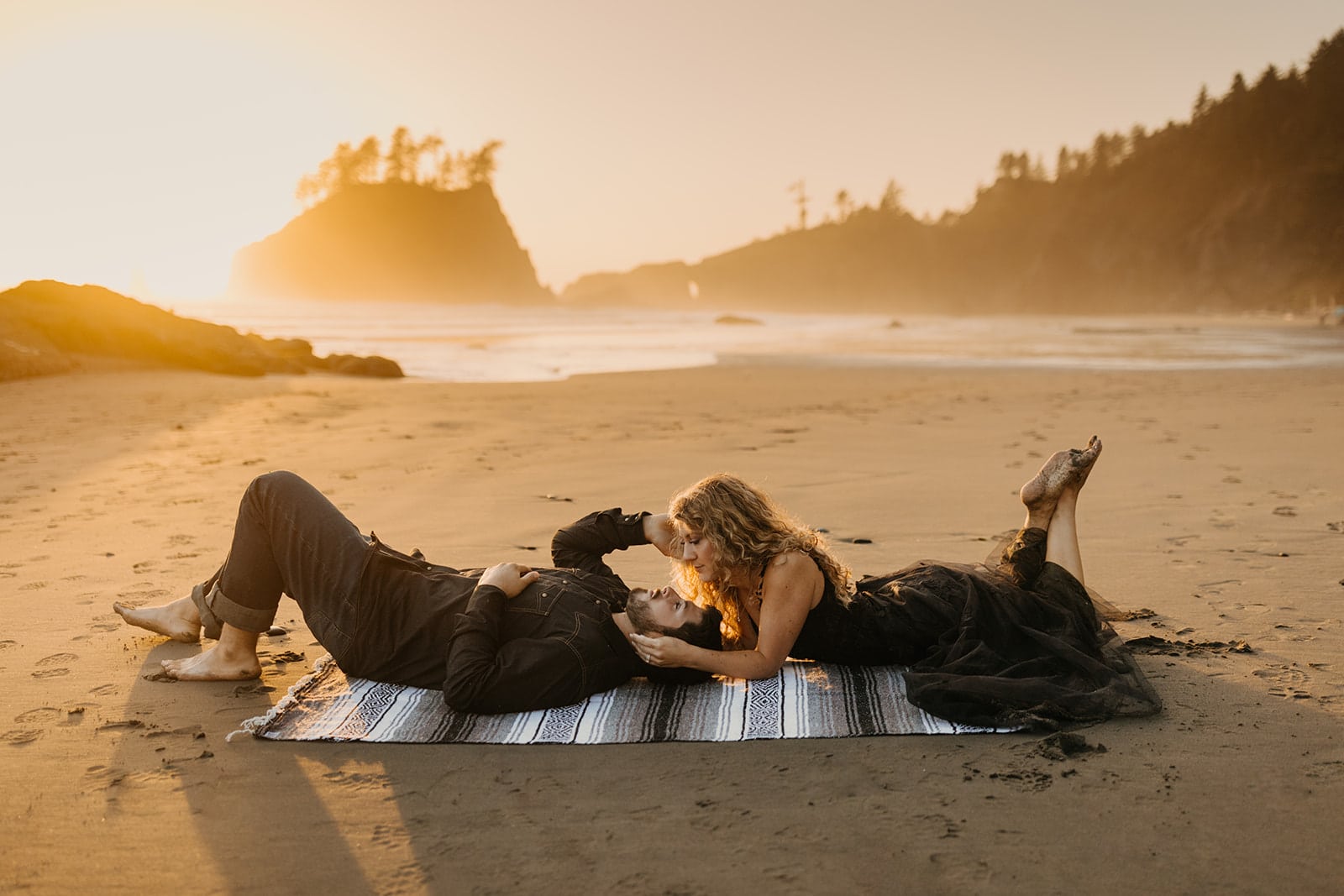 A bride wears a black wedding dress as she sits on the beach her husband.