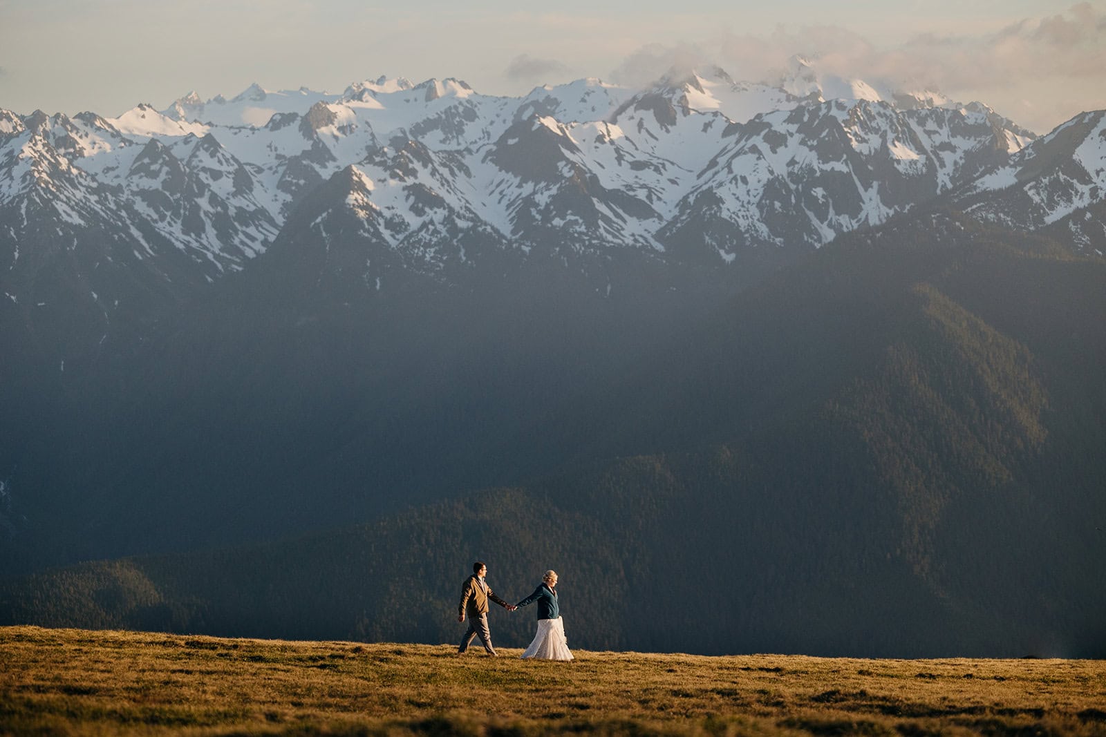 A bride leads her groom down the meadow trail with a massive mountain in the background.