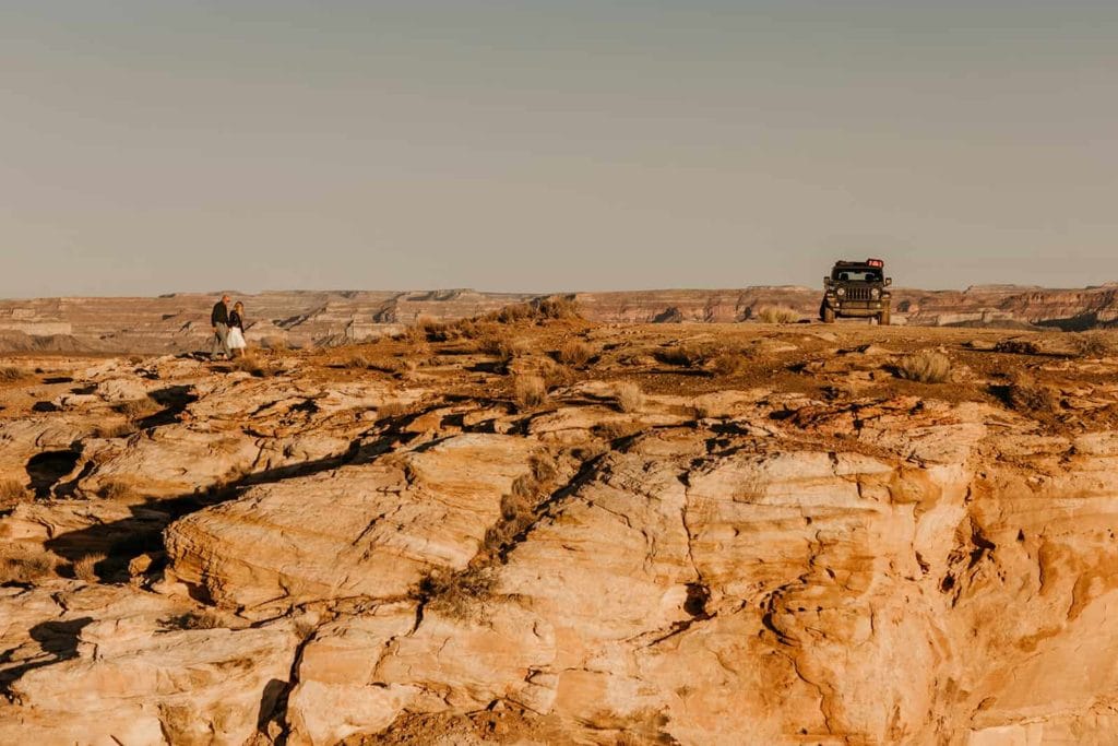 A couple stands together on a rock ledge with their jeep in the background