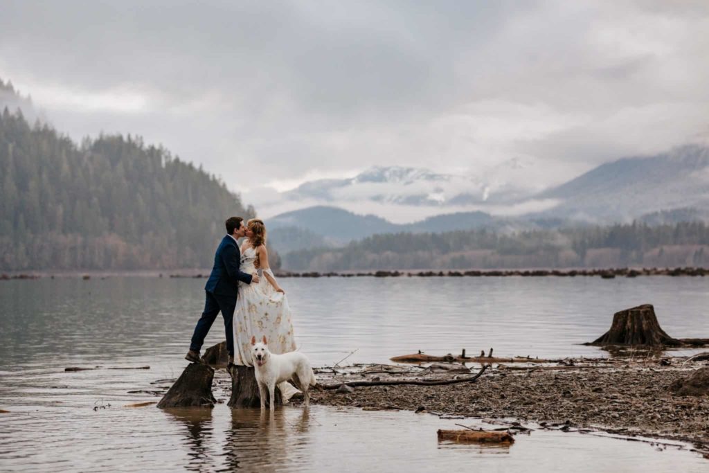 A couple kisses in the mountains as their dog looks at the camera.