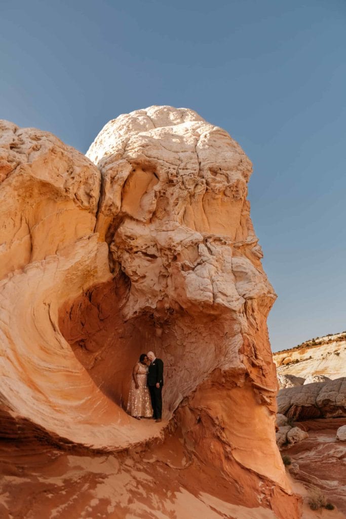 A couple stands within a red, orange and white rock in the desert.
