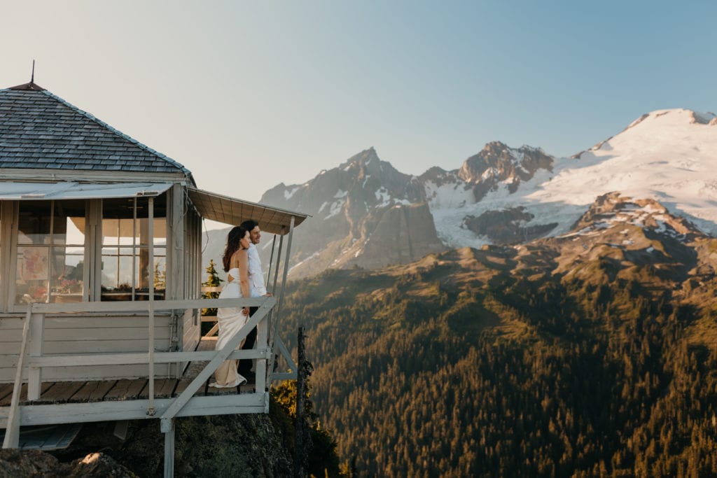 A bride and groom look out at the mountain view as they stand on a fire tower on their wedding day in the North Cascades.