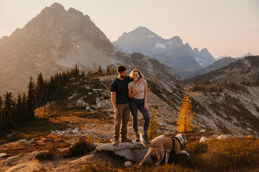 A couple smiles for a portrait with their dog in the mountains.
