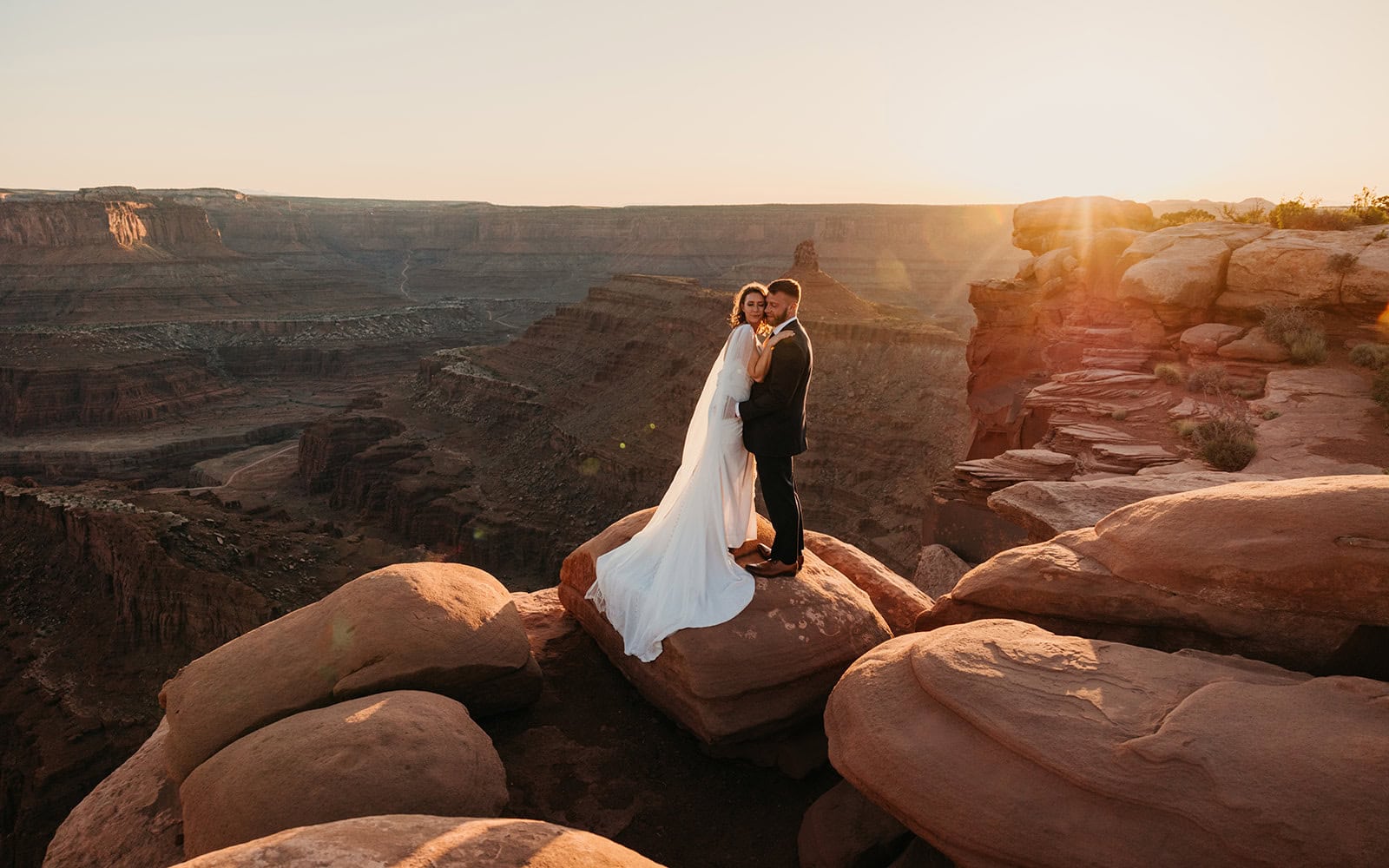 A couple stands on a ledge at sunset.
