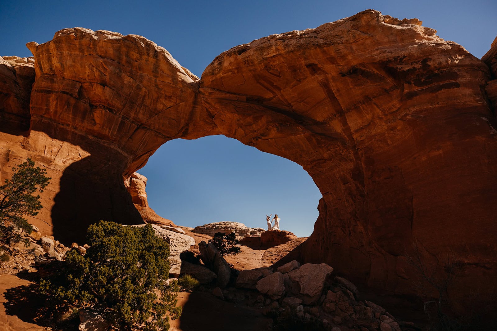 Two brides dance in the sunlight under a natural arch in arches national park.