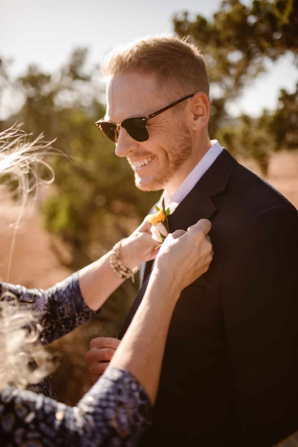 The groom smiling as his mom puts his flower arrangement on his suit.