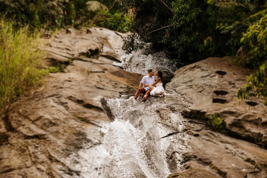 A couple hold each other as they sit in the water at a waterfall before sliding down together. 