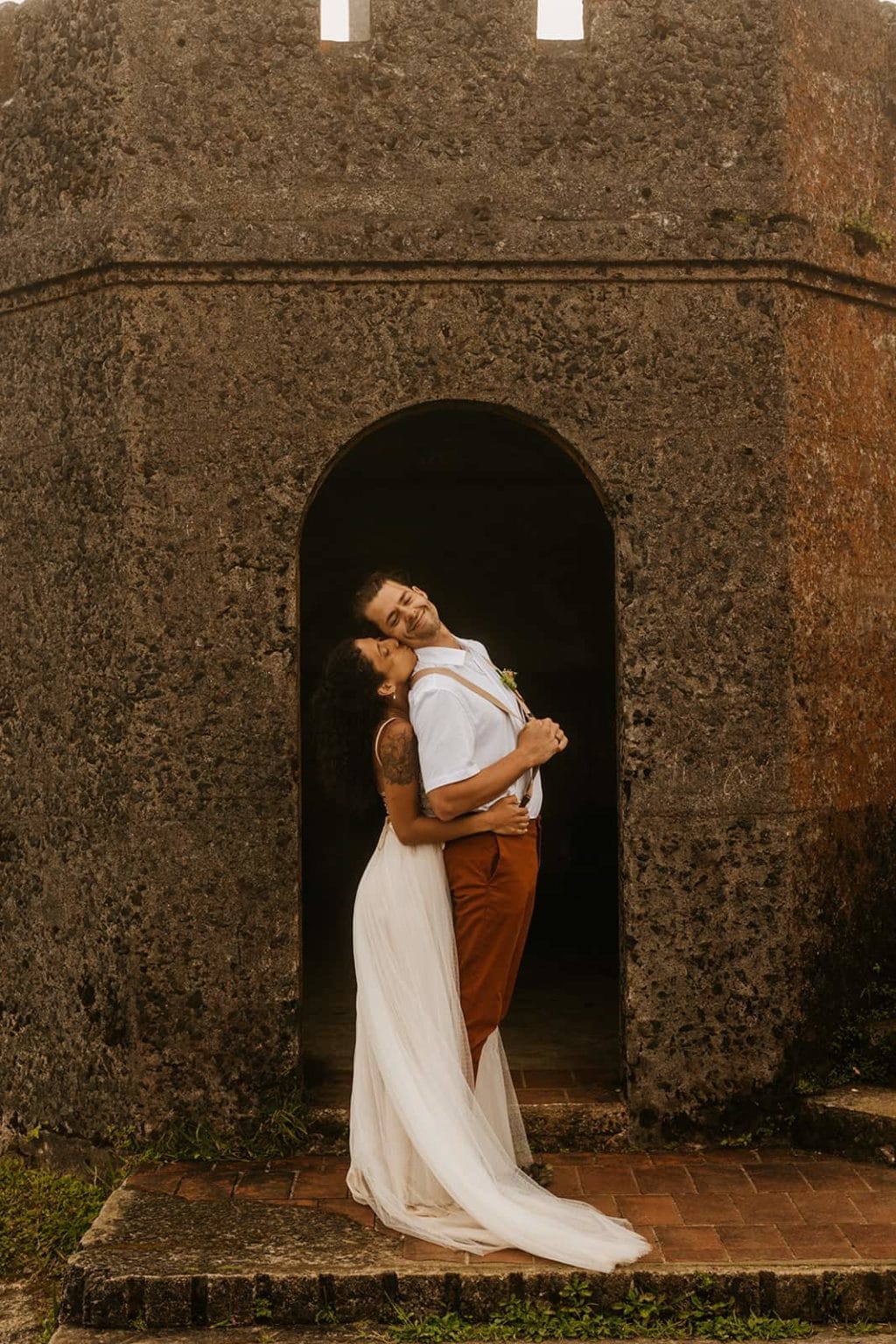 A groom smiles as his bride hugs and kisses him from behind.