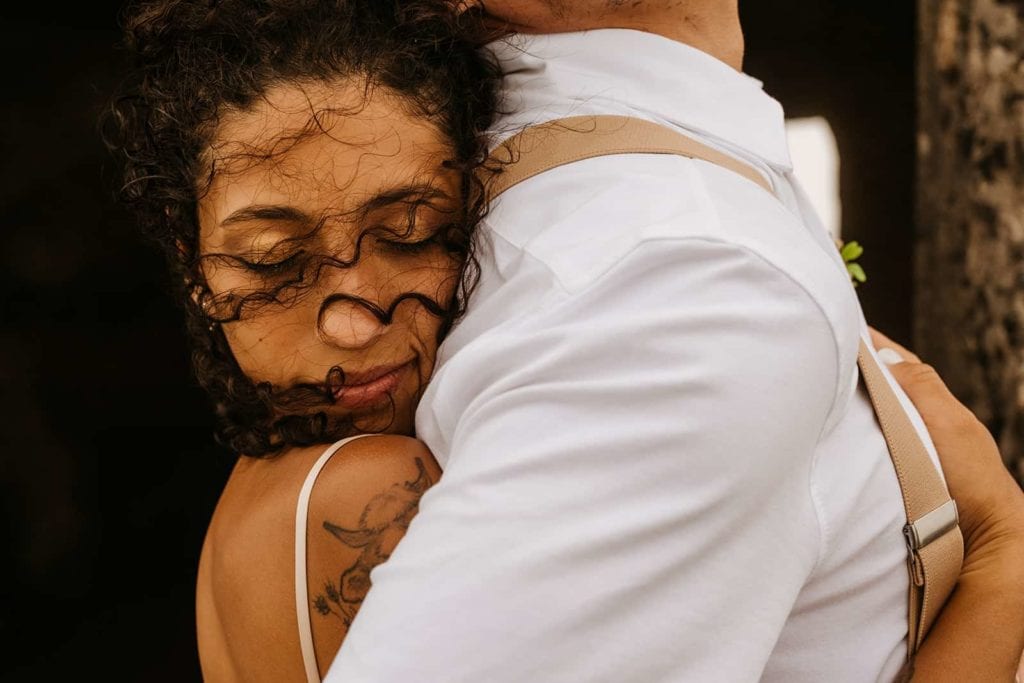 A bride embraces her groom from behind as the wind blows her fair in her face. 