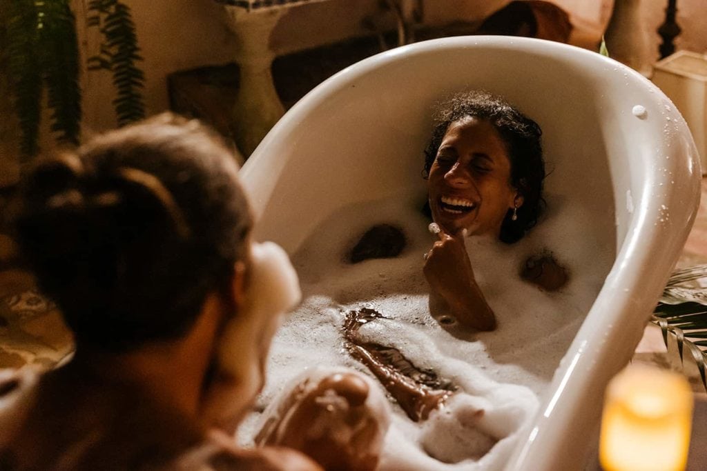 A woman laughs in the tub as her husband makes a beard out of soap suds. 