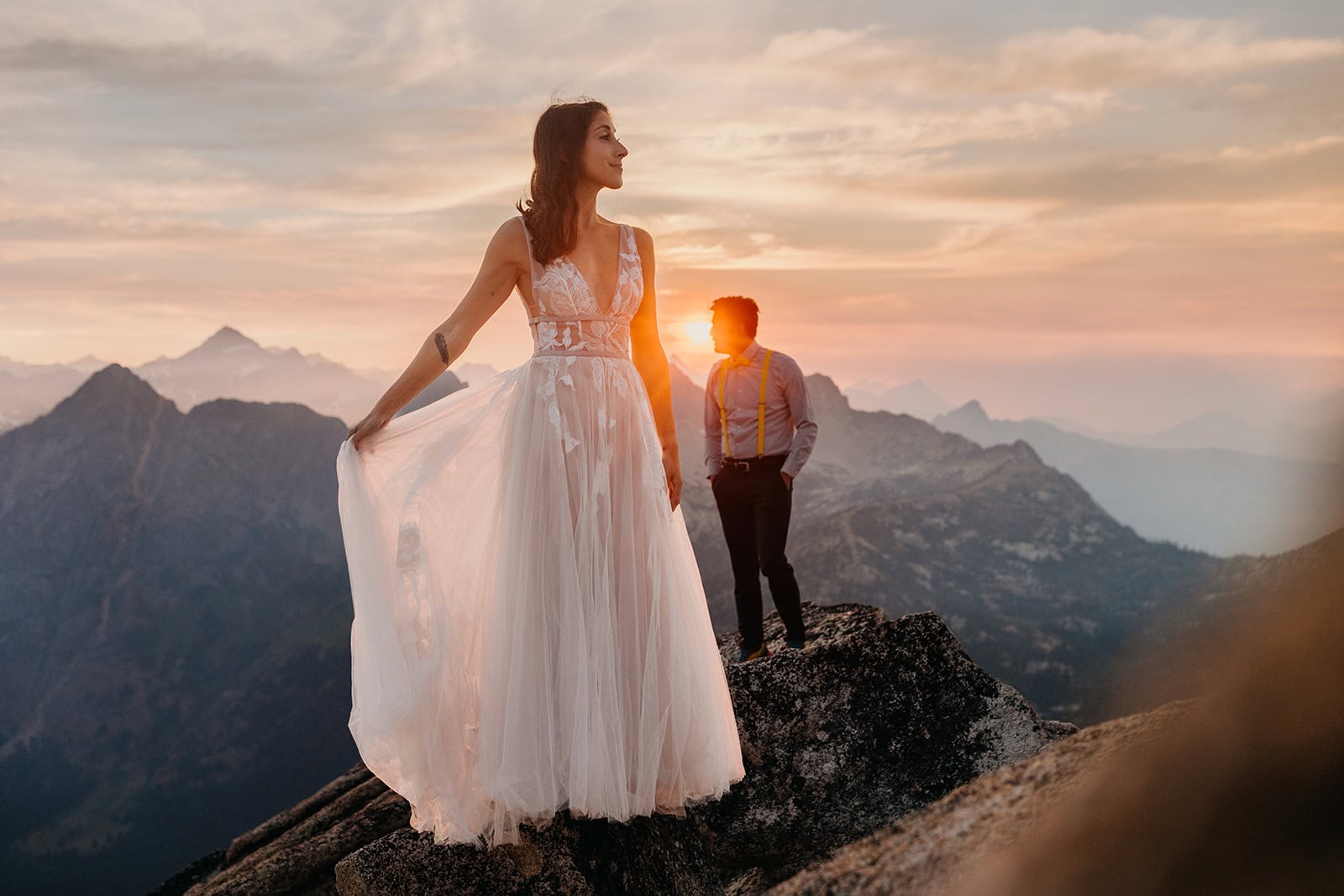A bride fans out her dress while her groom and the sunset are in the background