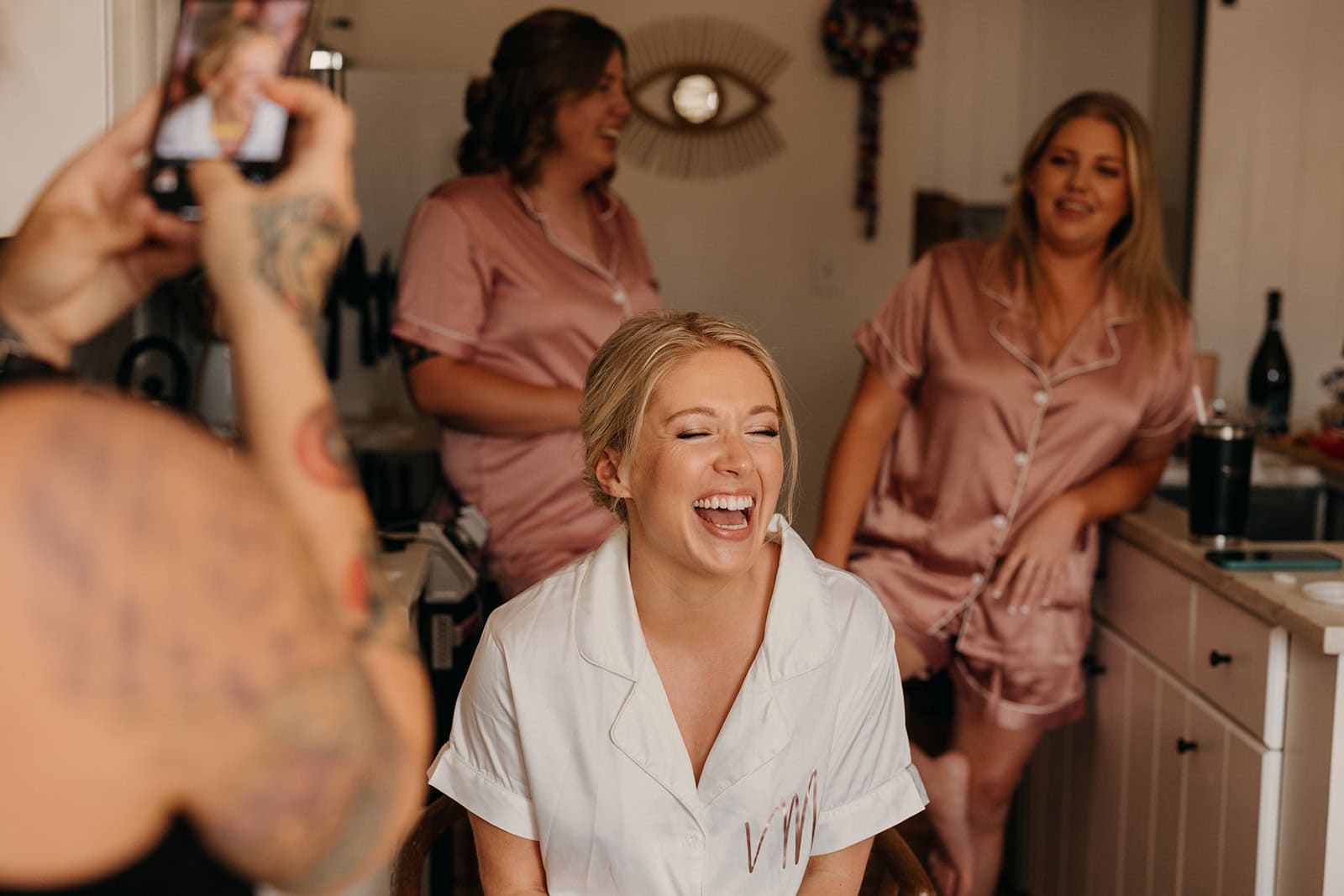 A bride laughs with her bridesmaids while doing hair and makeup.