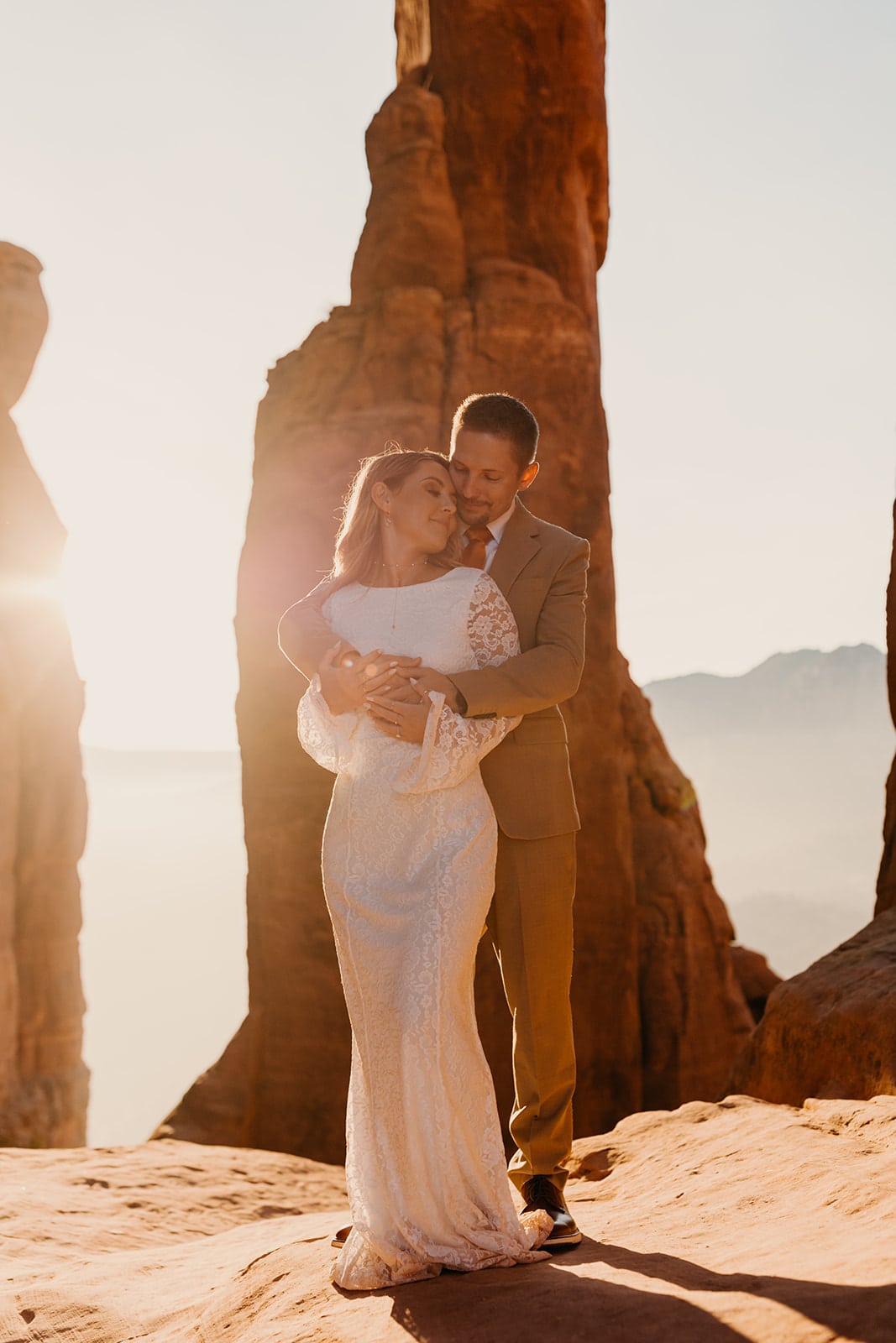 a man and his bride stand together in Sedona with the sunrise behind them between sandstone spires.