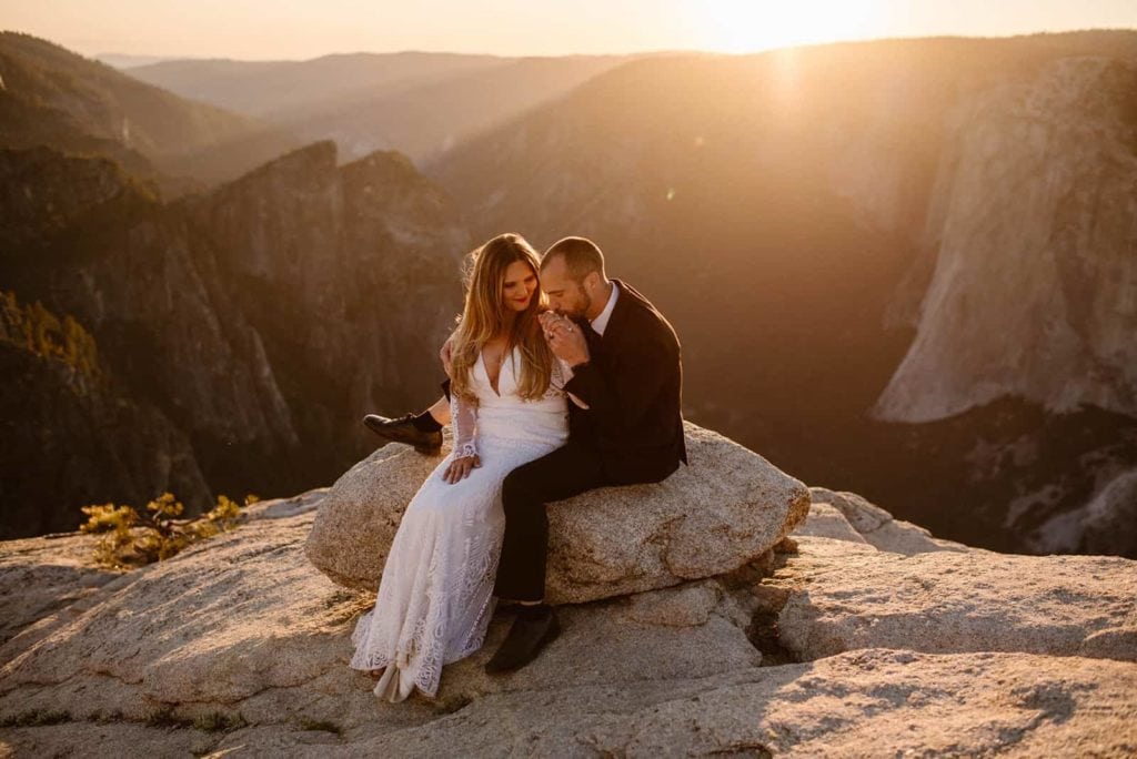 A groom kisses his wives hand as the sun starts to dip behind the big rock walls behind them as they sit enjoying sunset.