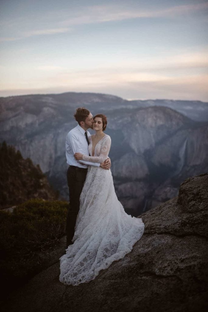 A groom kisses his bride on the check at dusk with a waterflow flowing behind them.